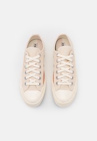 Baskets en toile beige avec un embout en caoutchouc blanc lisse, des panneaux latéraux contrastés, et des lacets plats blancs passés dans des œillets argentés.