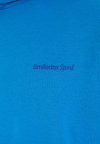 T-shirt sportiva blu realizzata in morbido cotone, caratterizzata da una superficie liscia con la scritta "Smilodox Sport" stampata in viola. Design semplice e funzionale.
