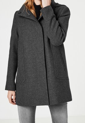 Kobieta w ciemnoszarym wełnianym płaszczu z wysokim kołnierzem i bocznymi kieszeniami, w połączeniu z jasnoszarymi dżinsami, na jednolitym tle.