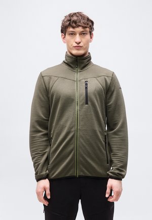 Nuori mies, joka käyttää oliivinvihreää, ribbineulottua vetoketjullista takkia, jossa on korkea kaulus ja mustat vetoketjutaskut, seisoo yksinkertaisen taustan edessä.