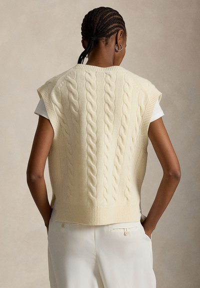 Osoba o zaplecionych włosach nosi kremowy bezrękawnik w warkoczowy wzór na białej koszuli i białych spodniach, stoi z rękami w kieszeniach.
