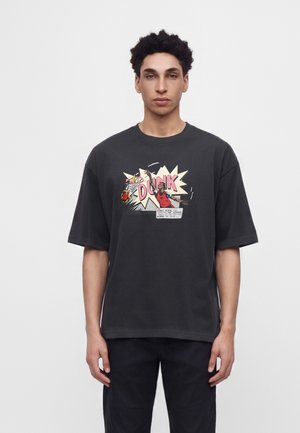 Jeune homme aux cheveux courts et bouclés porte un T-shirt noir oversize avec un motif coloré de dunk de basketball style bande dessinée et un texte à l'avant.