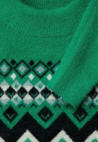 Pull vert en peluche avec un motif géométrique en diamants noir, blanc et gris, doté un col côtelé et une manche repliée sur la poitrine.