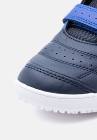 Κοντινή λήψη της μύτης ενός σκούρου μπλε αθλητικού παπουτσιού με διάτρητο δέρμα, μπλε λουράκι βέλκρο, μαύρη γλώσσα από πλέγμα και λευκή υφή σόλας σε λευκό φόντο.