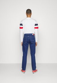Camicia bianca a maniche lunghe con accenti a strisce rosse, blu navy e rosse, abbinata a jeans in denim blu e sneakers rosa. Vista da dietro.