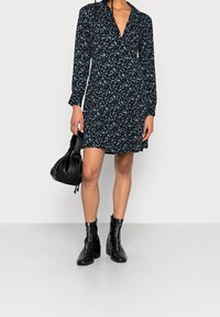 Kwiatowa sukienka z długim rękawem w czerni z różowymi i niebieskimi wzorami, zestawiona z czarnymi botkami do kostki i czarną torebką.