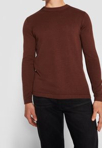 Pull en laine marron à manches longues avec un motif de tricot texturé et un col ras du cou, associé à un jean sombre. Aucune logo ou embellissement visible.
