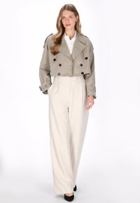 Gekürzter beiger Trenchcoat mit zwei Knöpfen, kombiniert mit hoch taillierter, weit geschnittener cremefarbener Hose und schwarzen spitz zulaufenden Schuhen.