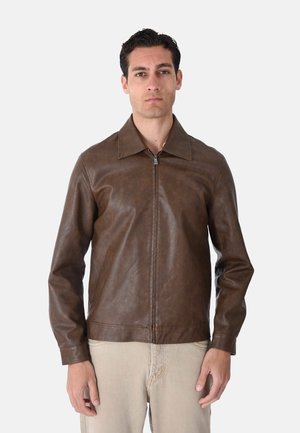 Uomo che indossa una giacca in pelle marrone con zip e ampio colletto e pantaloni beige, in piedi davanti a uno sfondo chiaro e uniforme.