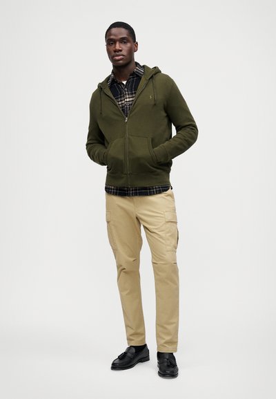 Casaco de capuz verde-oliveira com fecho à frente e um bolso na frente, sobre uma camisa de padrão checkered em preto e branco, combinado com calças cargo bege e sapatos pretos.