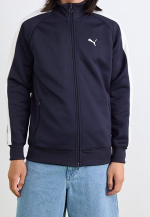 Person, die eine marineblaue Trainingsjacke mit Reißverschluss trägt, mit weißen Streifen an den Ärmeln und einem weißen Puma-Logo auf der Brust.