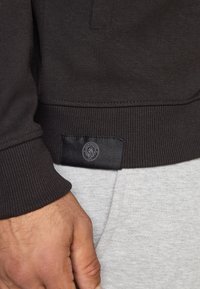 Ανδρικό χέρι κοντά στο στρίφωμα ενός μαύρου φούτερ με μικρή ετικέτα λογότυπου της Μάντσεστερ Σίτι π�άνω από ανοιχτού γκρι παντελόνι.