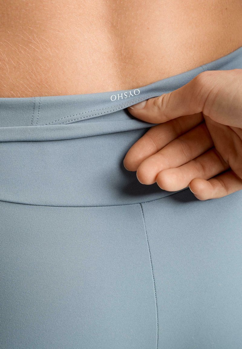 Svetlomodré športové legíny s hladkou textúrou, s pásom označeným logom. Ruka upravuje pás.