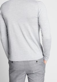 Grauer langärmeliger Strickpullover mit Rundhalsausschnitt, kombiniert mit grauen karierten Hosen, die über zwei Gesäßtaschen und Knöpfe verfügen.