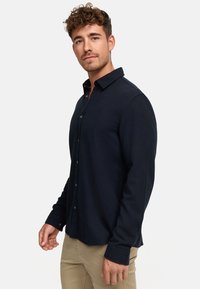 Camisa azul marino de botón con una textura suave, mangas largas y un cuello estándar; presenta un logo sutil en el pecho.