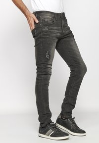 Slim-fit black denim jeans with distressed detailing and visible stitching, paired with black athletic sneakers featuring gray accents.