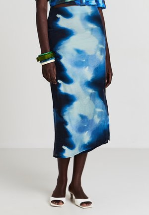 Jupe midi en tie-dye bleu et marine en tissu léger, présentant une coupe droite et un subtil éclat, associée à des chaussures ouvertes blanches.
