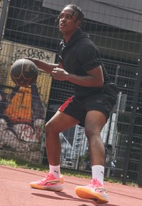 Basketballspiller i en sort hættetrøje og shorts med røde accenter, der dribler en bold, mens han har livlige pink og gule sneakers på en rød bane.