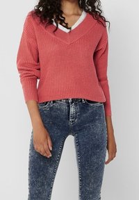 Roze gebreide trui met een V-hals, geribbelde textuur en lange mouwen, gecombineerd met een donkere skinny jeans.