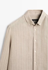 Beżowa lniana koszula z długim rękawem, delikatnym pionowym prążkowaniem, kołnierzykiem zapinanym na guziki i białymi guzikami na przodzie.