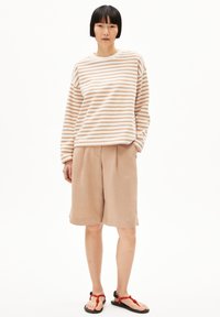 Gestreiftes Langarmshirt in Creme und Hellbraun, kombiniert mit beigefarbenen, plissierten Shorts und schwarzen Sandalen mit roten Akzenten.