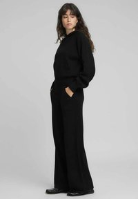 Sweat à capuche noir avec une poche frontale, associé à un pantalon noir ample. Le tissu semble doux avec une texture lisse. Design simple.