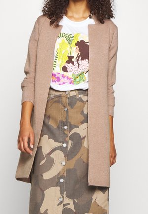 Vrouw draagt een beige openvallende cardigan, wit T-shirt met kleurrijke illustratie van plant en vrouw, en een rok met knopen en camouflagepatroon.