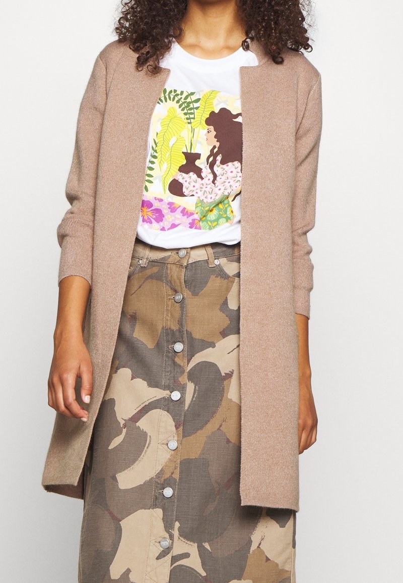 Femme portant un cardigan beige ouvert devant, un t-shirt blanc graphique avec une illustration colorée de plante et de femme, et une jupe boutonnière à motif camouflage.