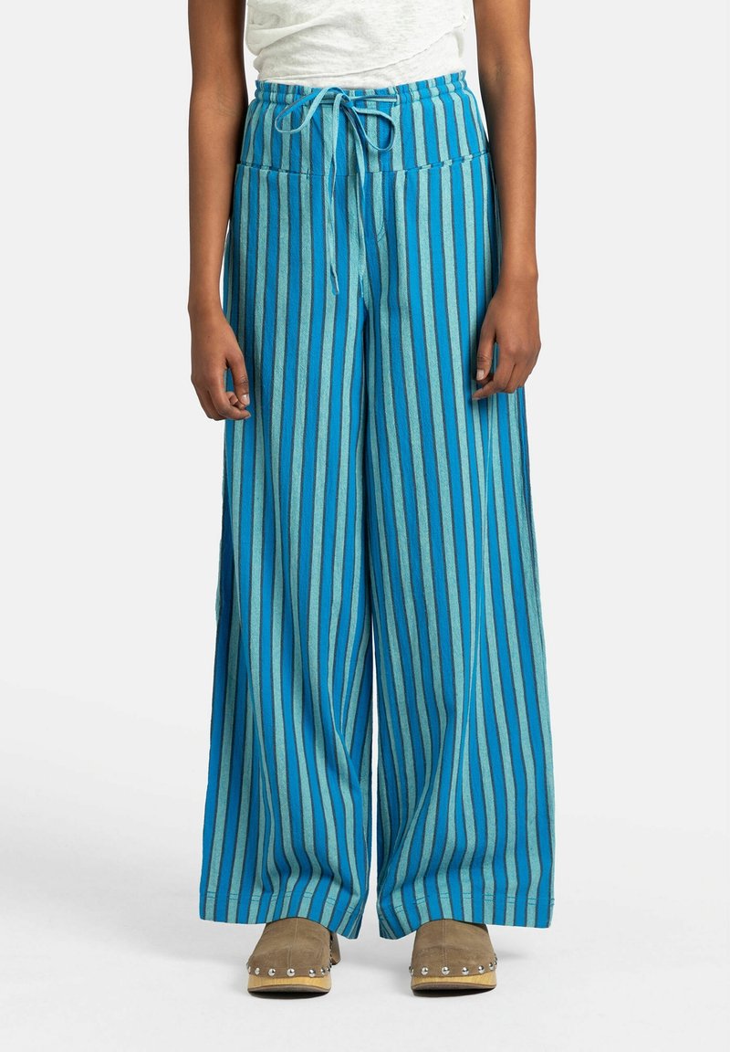 Breda byxor i blå och turkosa ränder. Har resår i midjan och ett mjukt, texturerat tyg. Kombineras med beige tofflor.