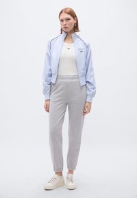 Gaiši zila bumbvedēja jaka ar rāvējslēdzēju, balta apaļas apkakles virsdrēbe, pelēkas sporta bikses, krēmkrāsas sporta apavi. Jakai ir aproces un izšūta logotipa detaļa.