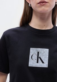 Μαύρο βαμβακερό t-shirt με ανάγλυφο γκρι τετράγωνο σχέδιο και τα γράμματα "cK" σε μαύρο. Κοντά μανίκια και στρογγυλή λαιμόκοψη.