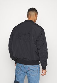 Svart bomberjacka med ribbade ärmar och nederkant. Med en texturerad tecknad karaktärsdesign på baksidan. Bärs över blå jeans.