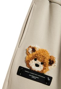 Bolsa de tela beige con un aplique de osito de peluche marrón, texturizado y suave. Etiqueta negra con texto blanco y dos botones negros adjuntos.