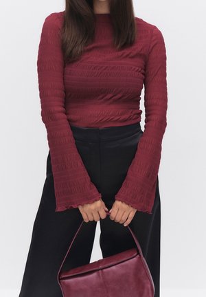 Kvinde iført en tekstureret langærmet bordeaux top og sorte bukser, der holder en matchende bordeaux håndtaske foran sig.