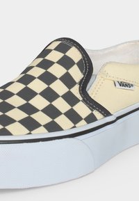 Chaussure Vans slip-on avec motif à damier noir et beige et semelle épaisse en caoutchouc blanc, montrant l'étiquette du logo Vans sur le côté en tissu beige.