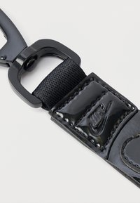 Juodas blizgus raktų pakabukas su tekstūruota dirželiu, puoštas siūlėmis, ir metaliniu juodu užsegimu. Nike logotipas įspaustas ant dirželio.