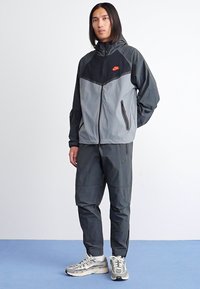 Nike Trainingsjacke und -hose in Schwarz und Grau, aus leichtem Material, mit Reißverschlusstaschen, mit Logo auf der Brust, kombiniert mit grauen Sneakern.