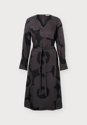 Femme portant une robe noire et grise à fleurs, longueur genoux, manches longues avec ceinture à la taille, associée à des talons noirs pointus.