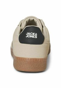 Sneaker beige avec une tige en suède texturé, un onglet de talon en cuir noir arborant un logo blanc, et une semelle extérieure en caoutchouc gomme avec un motif antidérapant.