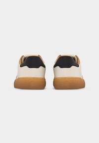 Ζευγάρι χαμηλά αθλητικά παπούτσια με λευκό επάνω μέρος, μαύρη ετικέτα στη φτέρνα και χοντρές καφέ λαστιχένιες σόλες, που φαίνονται από πίσω σε λευκό φόντο.