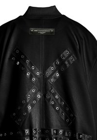 Chaqueta negra con un alto cuello, etiqueta de cuero y correas decorativas con ojales en un patrón X en la parte posterior. Textura suave.