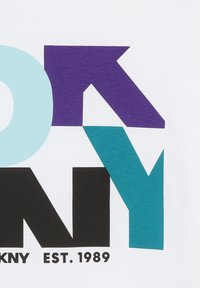 T-shirt en coton blanc avec des lettres géométriques audacieuses en bleu clair, violet, noir et turquoise, avec "EST. 1989" imprimé en dessous.