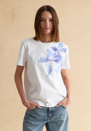 Mujer joven de pie con las manos en los bolsillos de los vaqueros, vistiendo una camiseta blanca con un gran estampado floral azul en el frente.