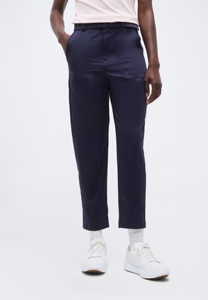 Person trägt marineblaue gerade geschnittene Hosen, weiße Socken und weiße Turnschuhe, steht mit den Händen in den Taschen vor einem schlichten Hintergrund.