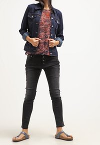 Chaqueta de mezclilla con lavado oscuro, mangas azules y cierre de botones sobre una blusa de estampado paisley rojo. Jeans skinny negros y chanclas marrones completan el look.