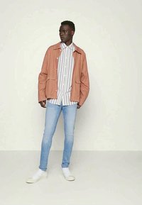 Casaco rosa claro com dois bolsos frontais, vestido sobre uma camisa riscada em branco e cinza. Jeans azul claro e ténis brancos completam o visual.
