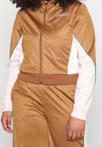 Person wearing brown Adidas track jacket and pants with light pink sleeves and green trim, standing with arms relaxed by sides.