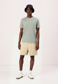 Lys grøn bomulds t-shirt, beige shorts, hvide sneakers med beige detaljer og en sort bøttehat. Enkel, afslappet pasform og minimale detaljer.