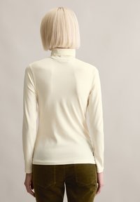 Langärmliges, cremefarbenes Rollkragenshirt mit figurbetontem Schnitt und weicher Textur, kombiniert mit grünen Cordhosen mit Gesäßtaschen.