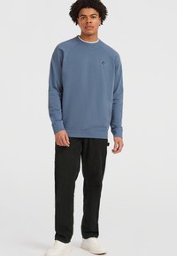 Blå crewneck sweatshirt med raglanærmer, ribbede manchetter og en subtil logoaccent på brystet, parret med sorte bukser og hvide sko.